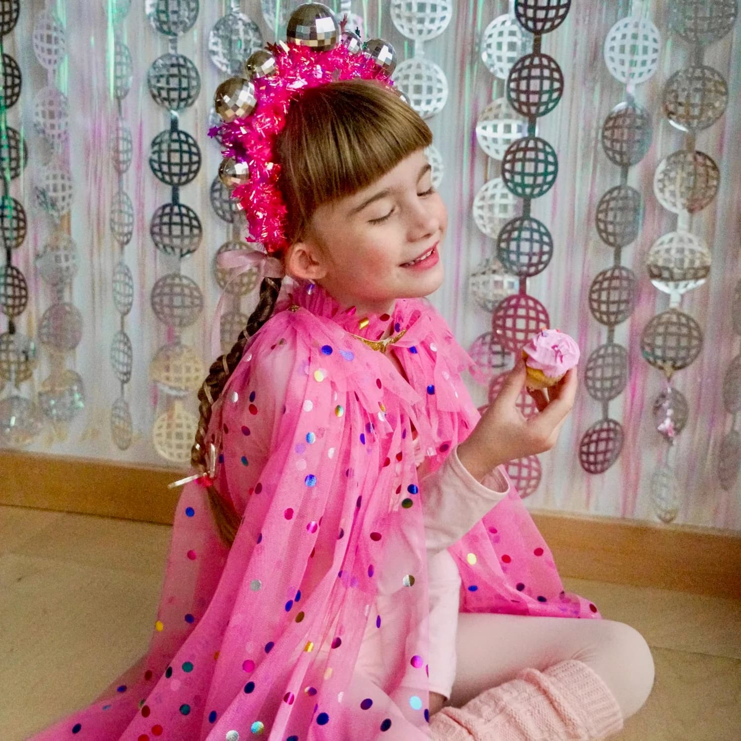 A smiling girl in a pink polka-dot cape and a disco-ball crown holding a frosted cupcake